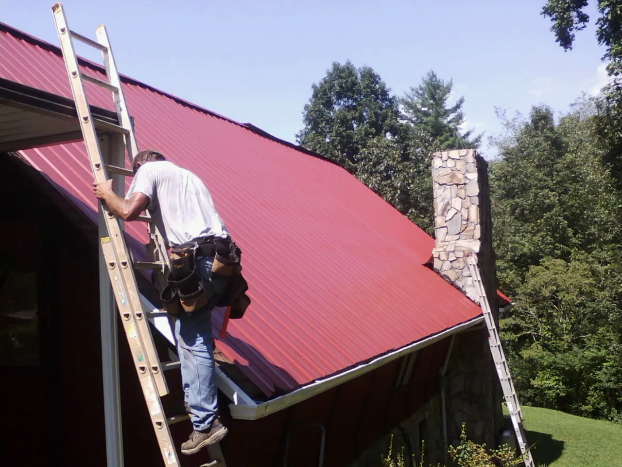 Roofer on a ladder beside a red metal roof and stone chimney — Soffit & Fascia Repair by Roofing Brothers in Stevinson